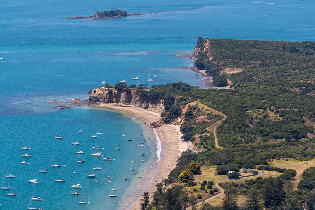 aerial shot of the coast of Waiheke Island covered in grass and green trees with boats in the blue ocean below - Top Snowbird Destinations For Canadians to Escape the Winter Chill new-zealand-auckland-waiheke-island-coastline TA